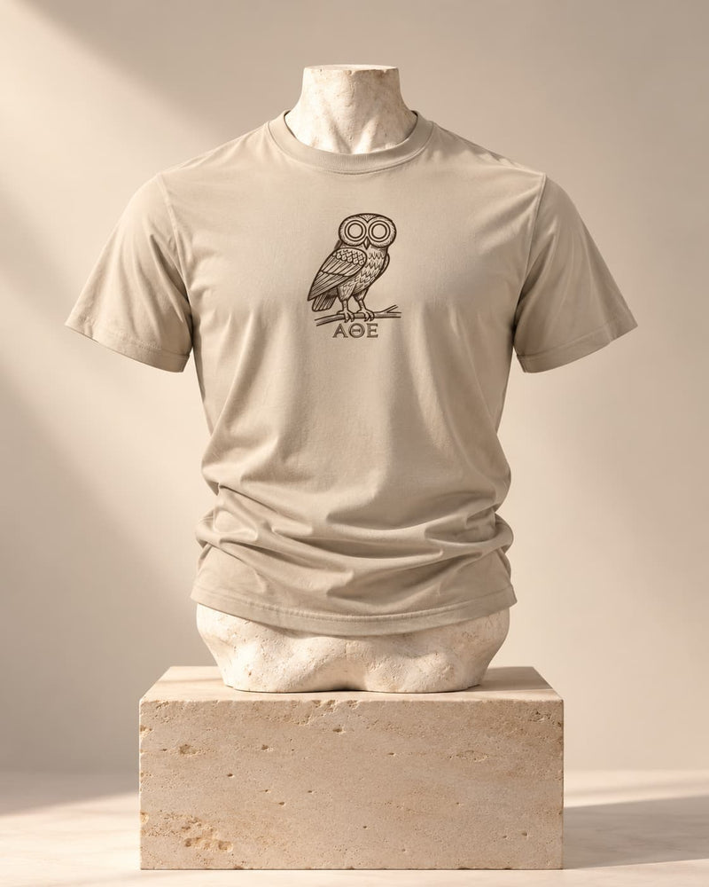 Camiseta ΑΘΗΝΑΙΩΝ en arena, sobre un busto de piedra y un pedestal de travertino, con una lechuza de trazo lineal y la inscripción «ΑΘΕ»